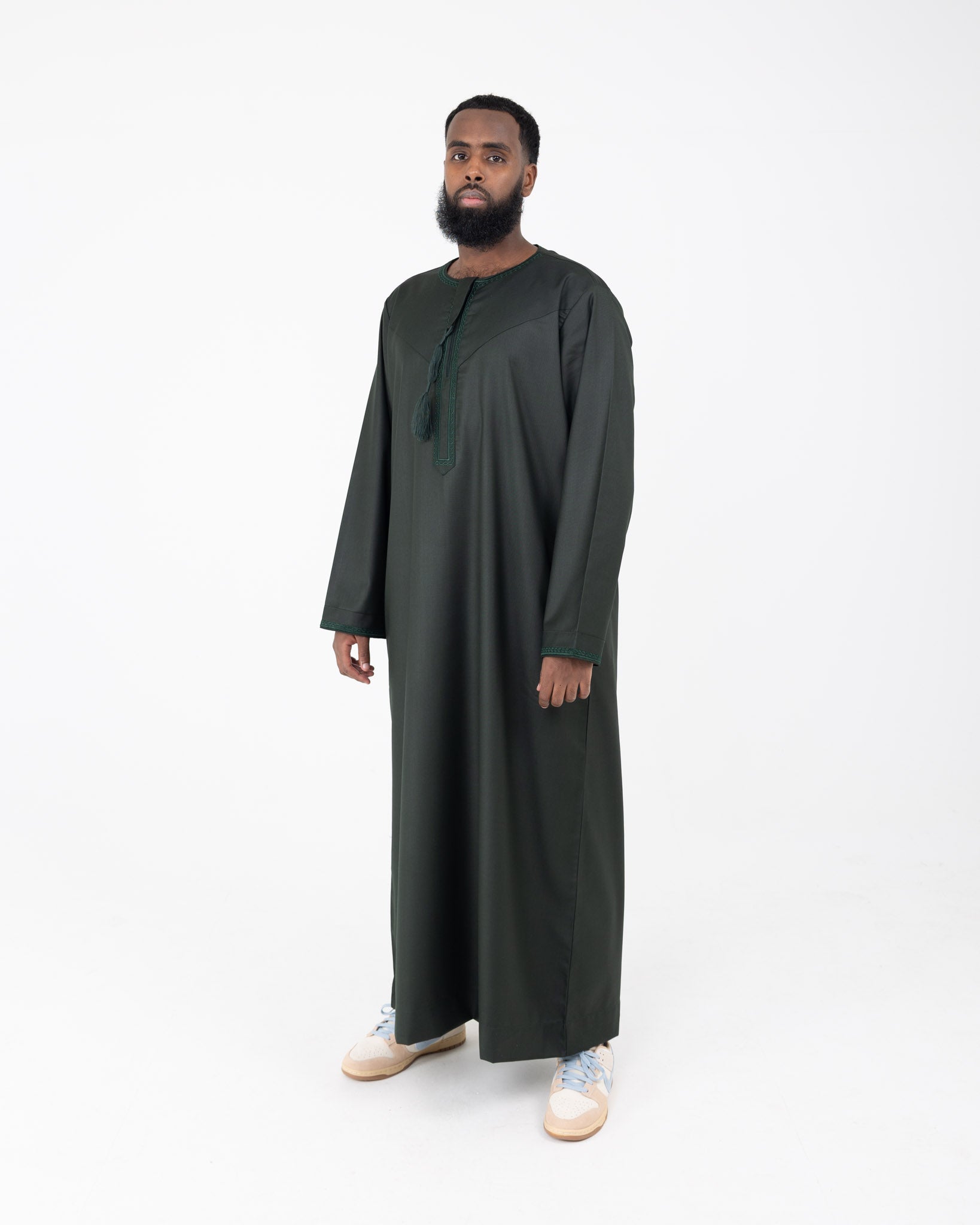 A man standing against a white background wearing an embroidered Rayan green thobe with embroidery on the neckline and cuffs.