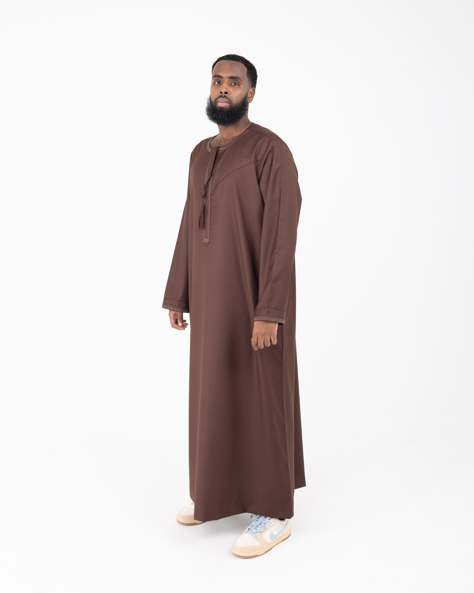 A man wearing a brown Rayan thobe with embroidery on the neckline and cuffs.