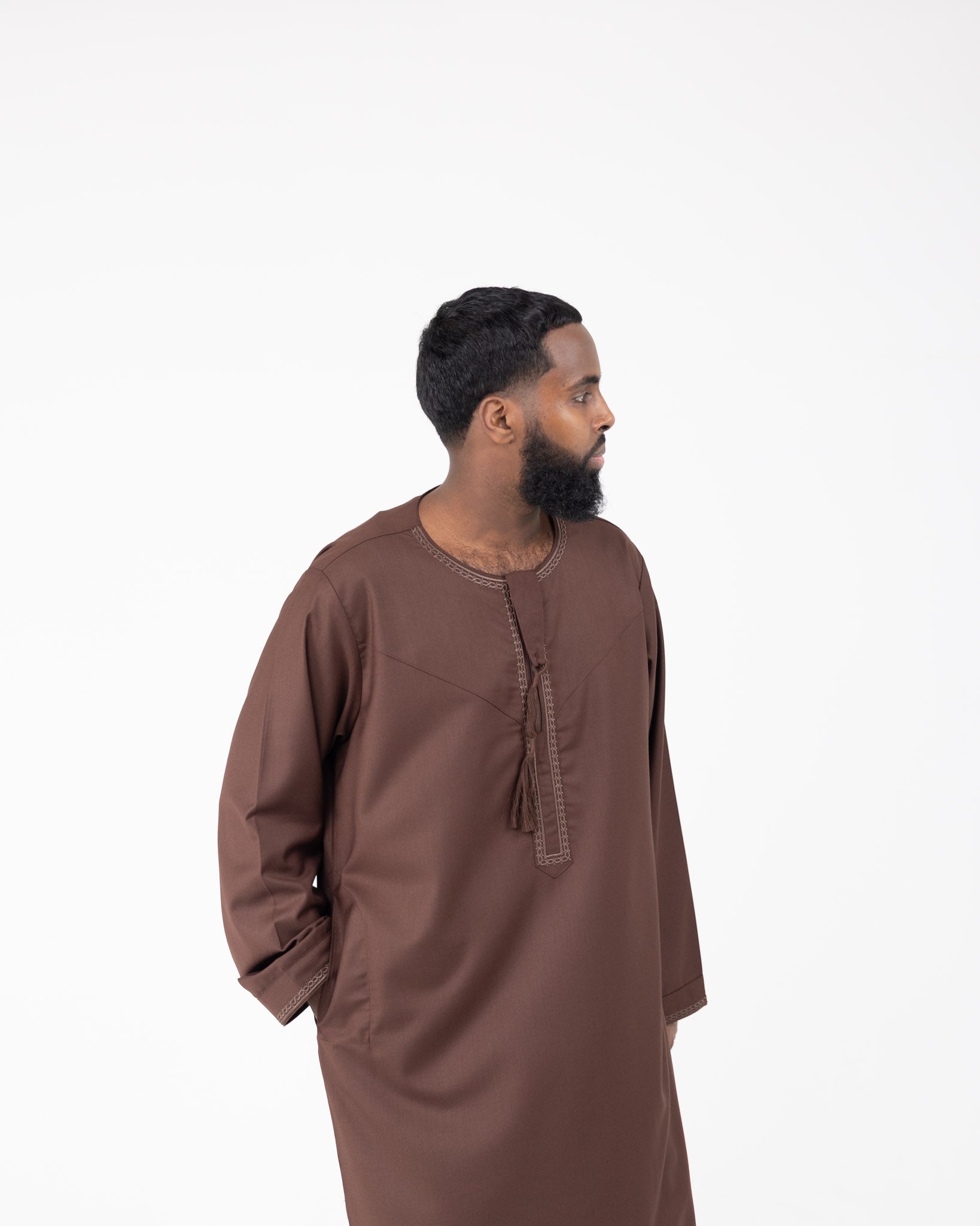 A man wearing a brown Rayan thobe with embroidery on the neckline and cuffs.