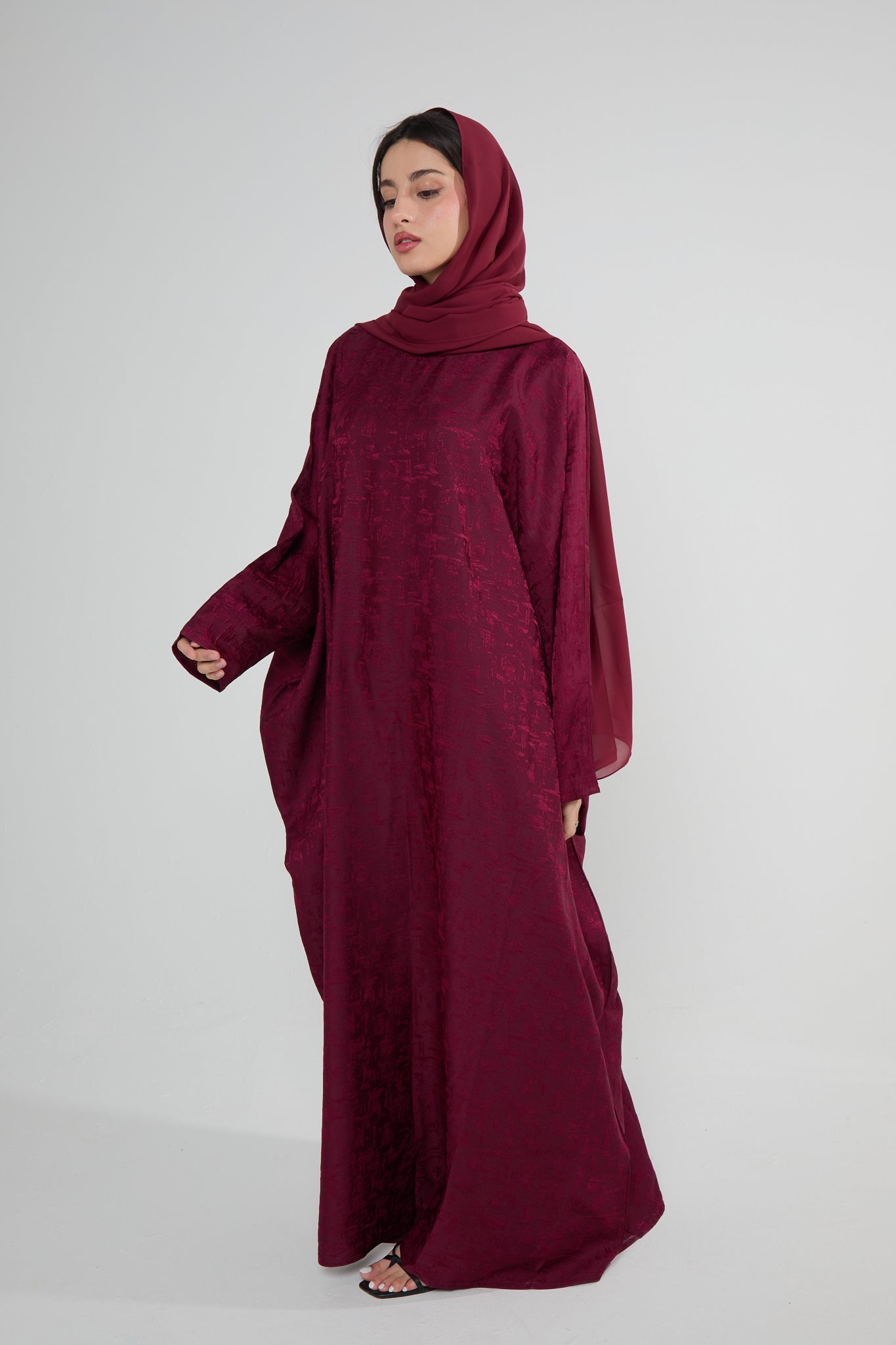 A woman wearing a maroon butterfly-cut abaya with a matching headscarf, standing against a plain background.