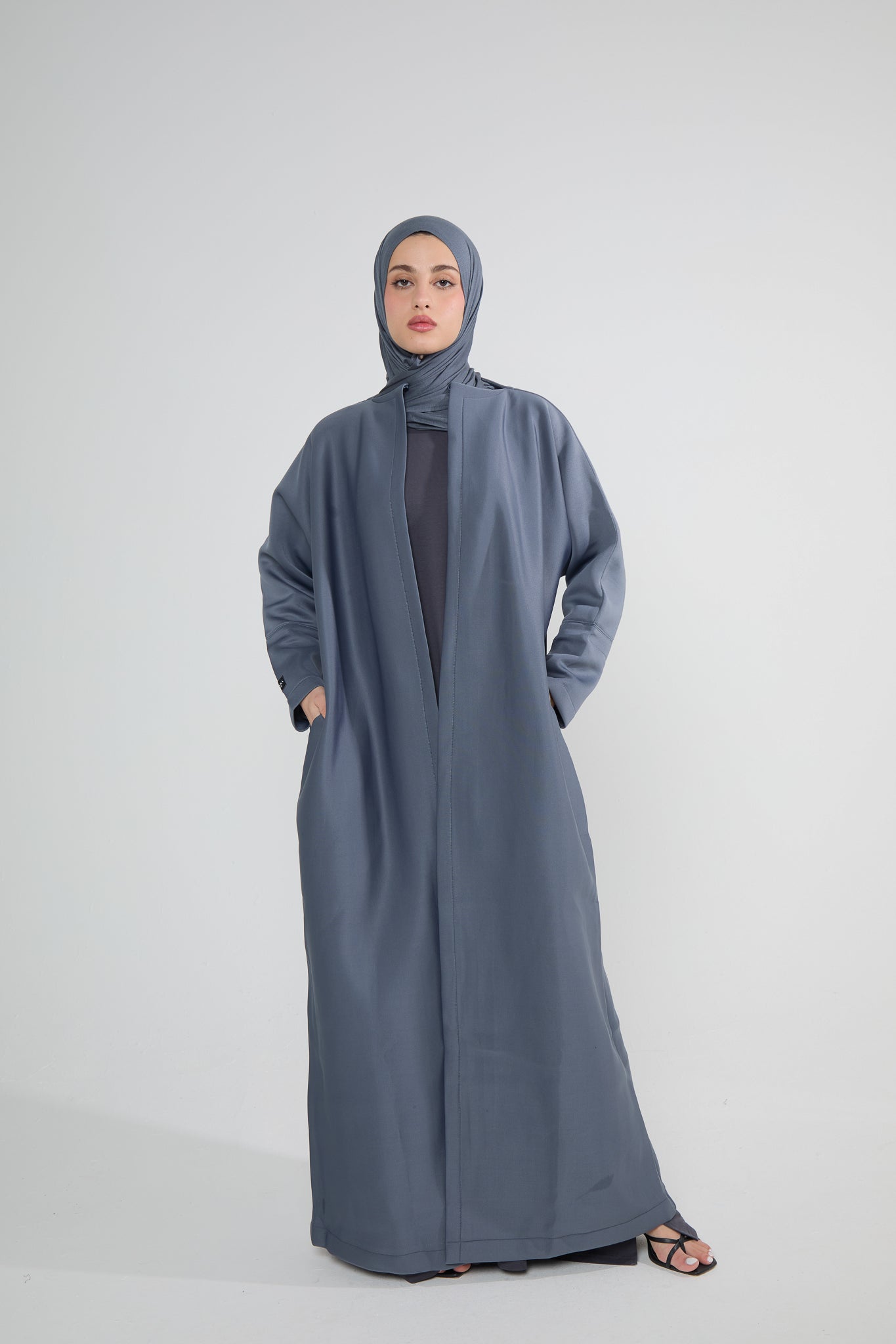 A woman wearing a grey neoprene abaya with an open wrap design, discreet hook closure, and functional side pockets.