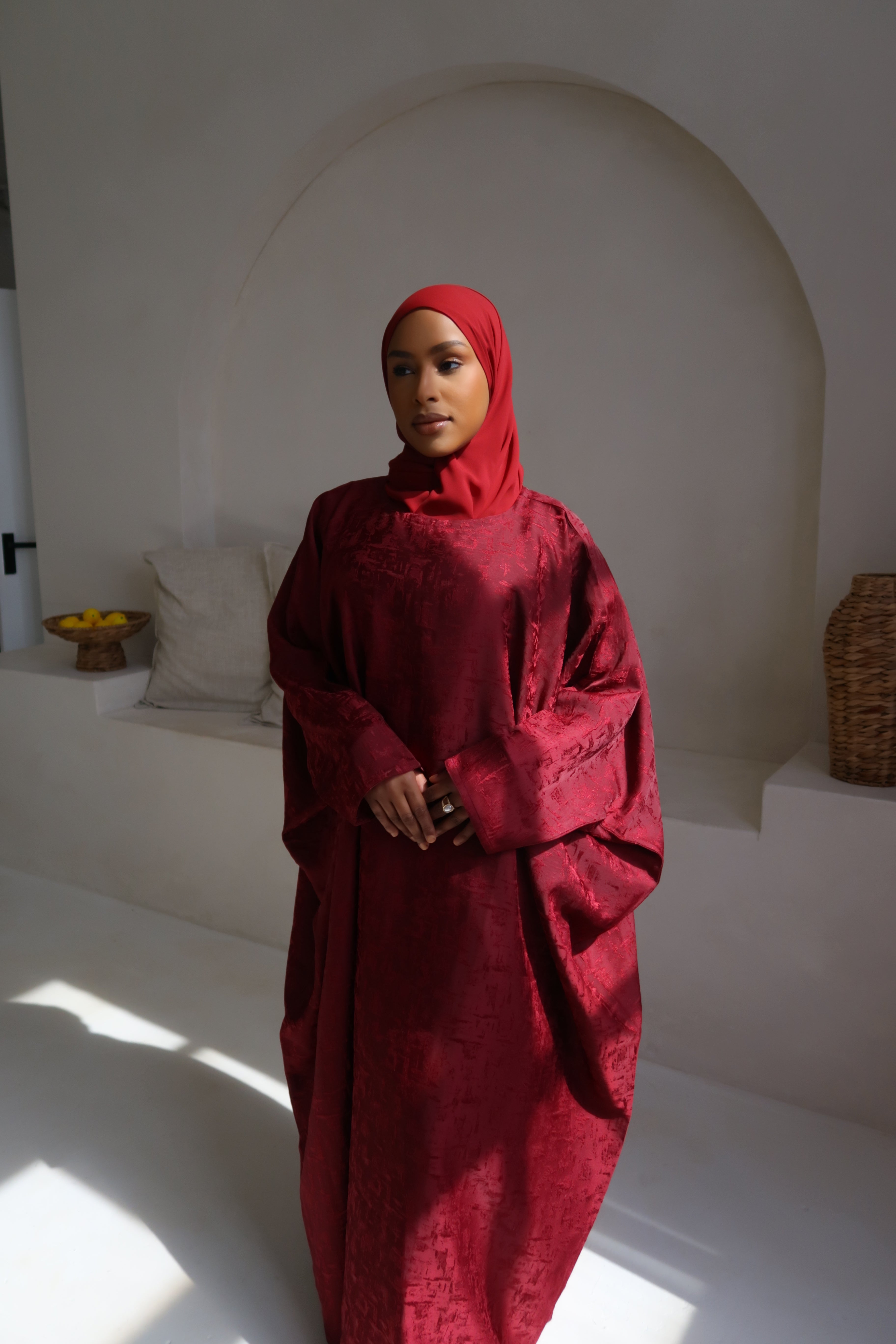 A model standing in a room wearing a red butterfly abaya with a matching headscarf, against a white background with arches and a window.