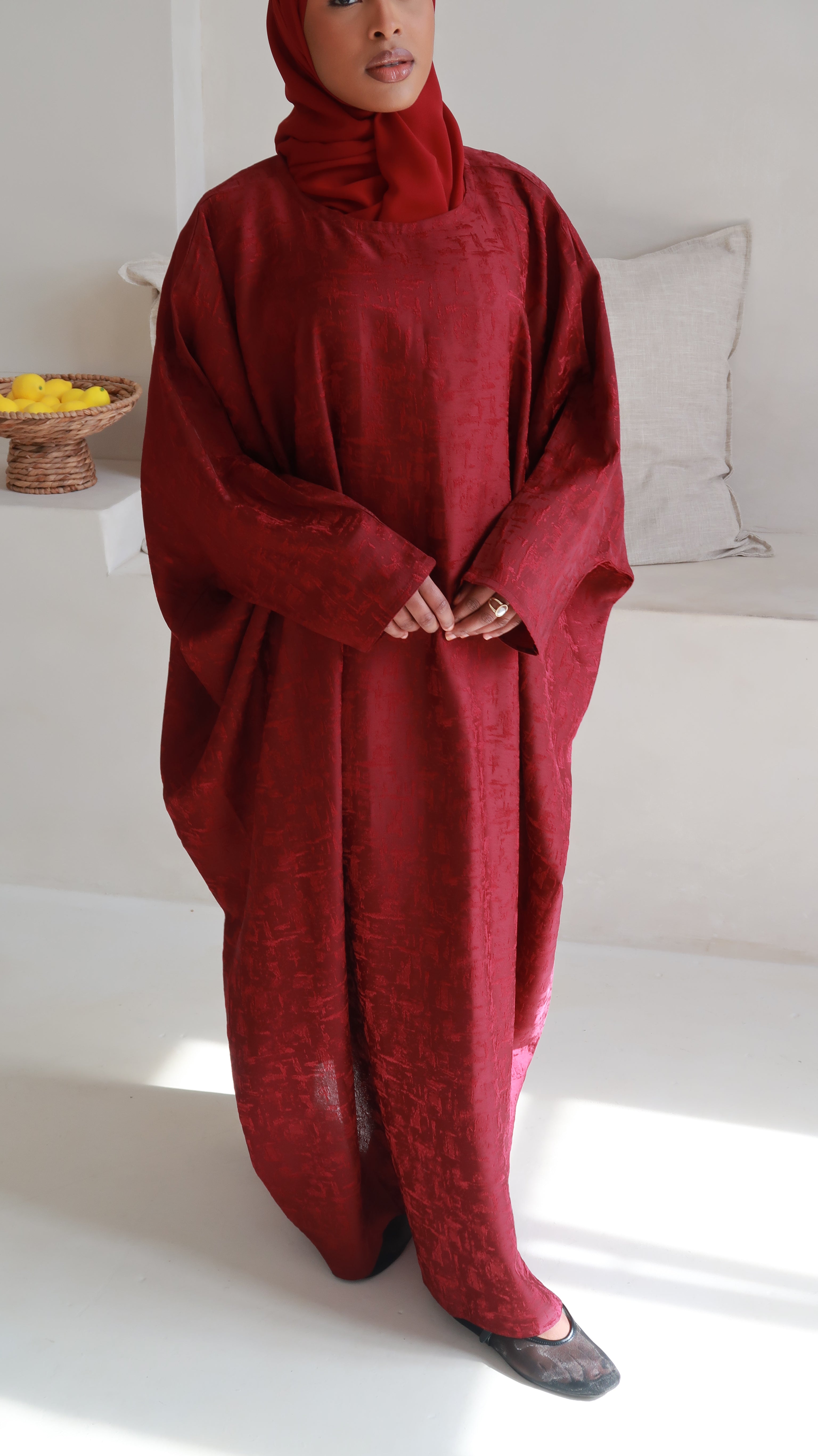 Close up of model standing in a room wearing a red butterfly abaya with a matching headscarf, against a white background with arches and a window.