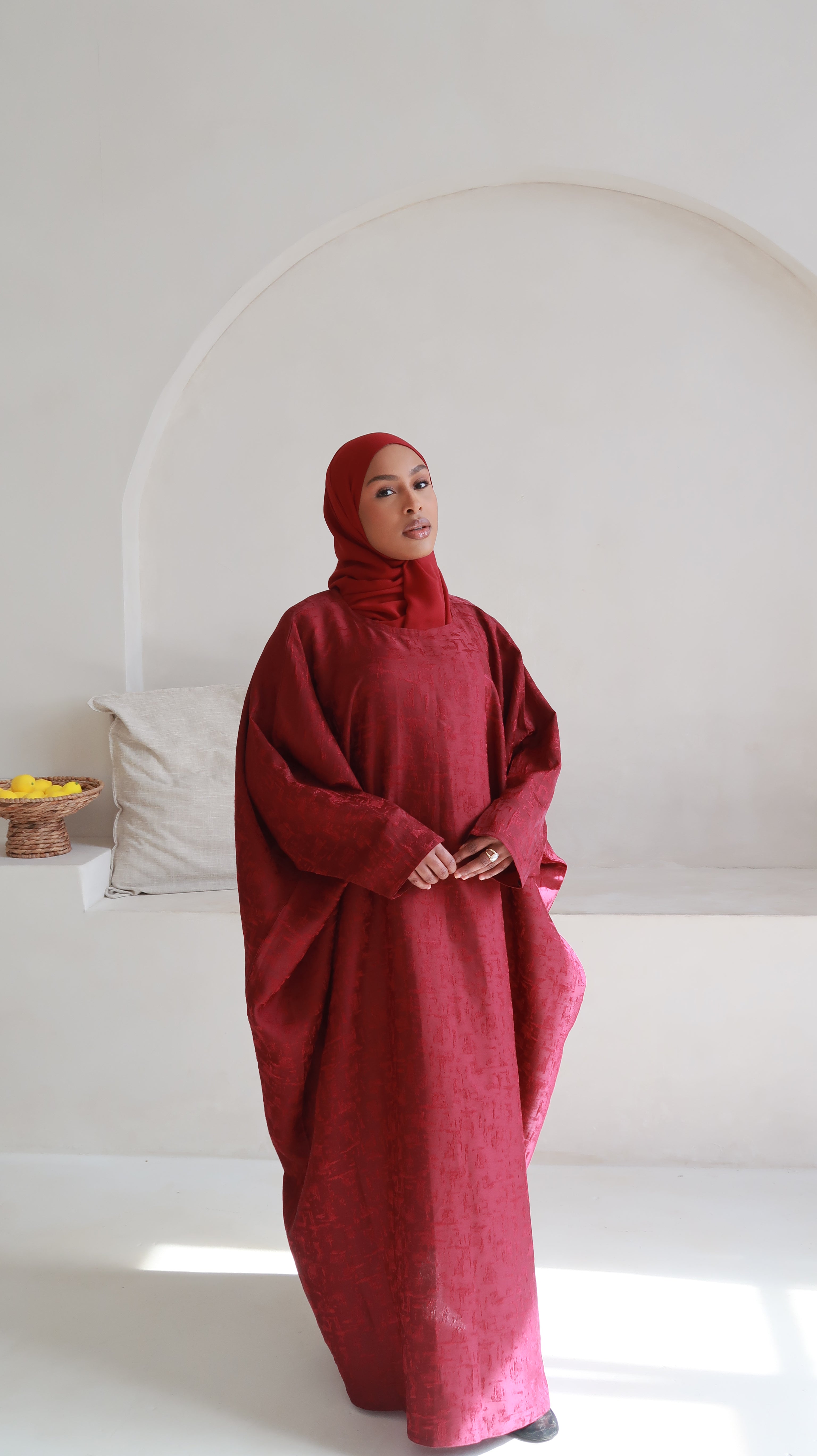 A model standing in a room wearing a red butterfly abaya with a matching headscarf, against a white background with arches and a window.